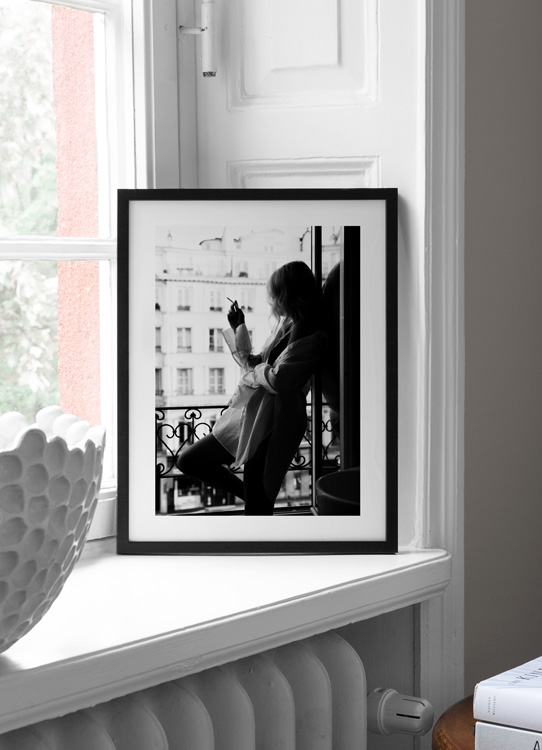  – Fotografia in bianco e nero di una donna che indossa una camicia oversize e fuma una sigaretta in piedi a una finestra